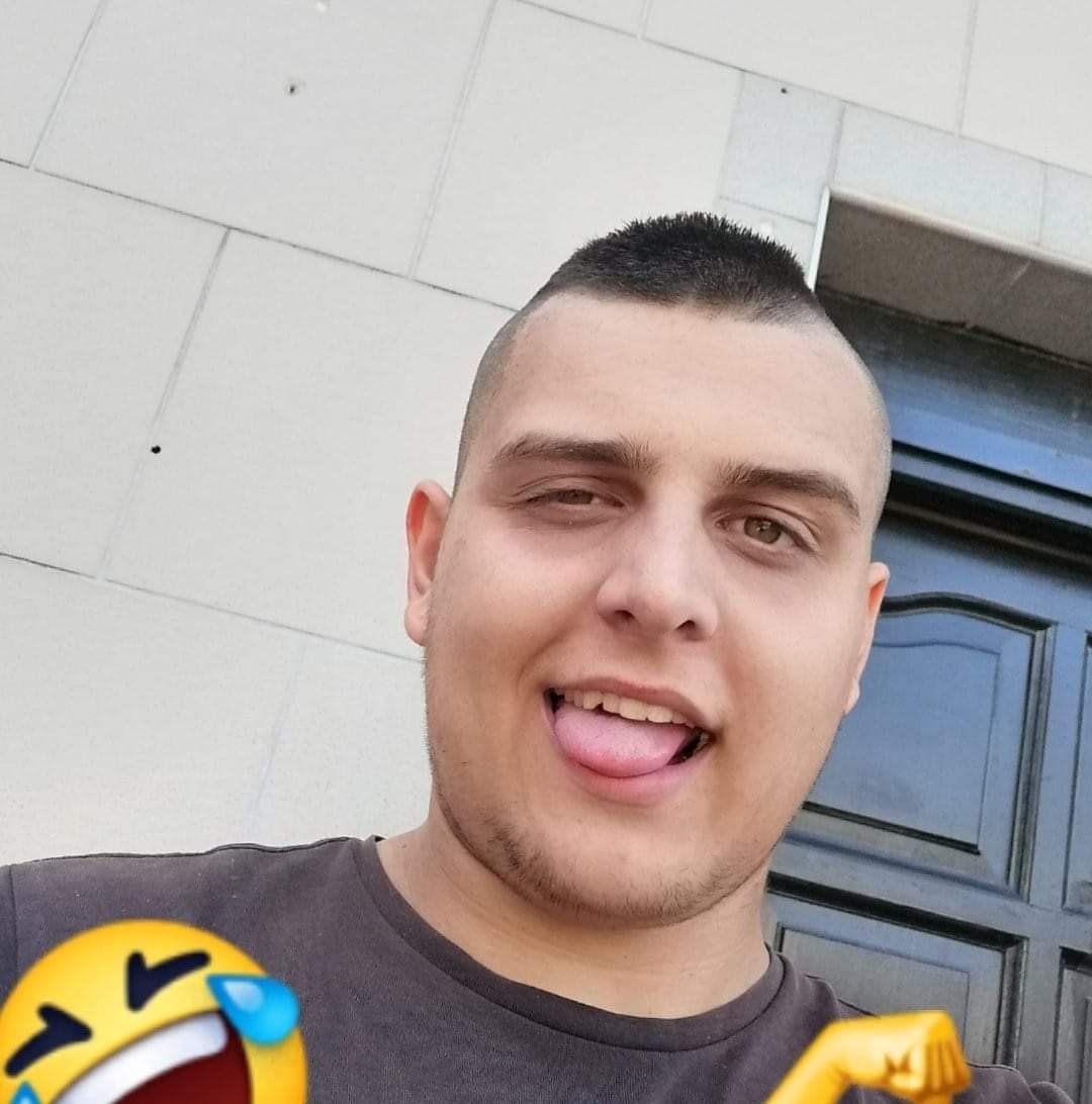 Portret mężczyzny z krótką fryzurą, wystawiającego język, z emoji w rogach zdjęcia, na tle jasnej ściany i ciemnych drzwi.