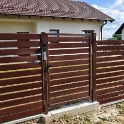 Brązowa, pozioma brama wjazdowa z domofonem, zamontowana na betonowej podmurówce, na tle jasnego domu z brązowym dachem.