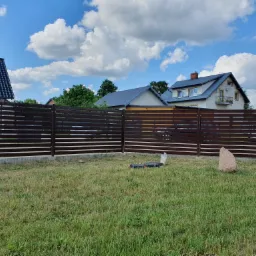 Nowoczesne ogrodzenie z poziomych paneli w kolorze ciemnego brązu, widoczne na tle domów i błękitnego nieba z obłokami. Na trawniku przed ogrodzeniem leżą narzędzia i stoi duży kamień.