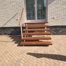 Zewnętrzne schody z drewnianymi stopniami i białą metalową konstrukcją prowadzące do drzwi, na tle ceglanej ściany budynku.