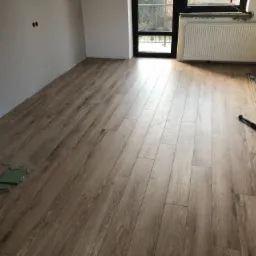 Nowo ułożone panele podłogowe w jasnym odcieniu drewna, widoczne narzędzia i akcesoria montażowe, ściany w szarym odcieniu, okno z widokiem na zieleń.