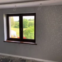 Ściana pokryta szarą tapetą ze wzorem, widok na okno z brązową ramą i parapetem, przez które widać zieleń drzew i żółty pojazd, na suficie widoczne wystające kable po lampie.