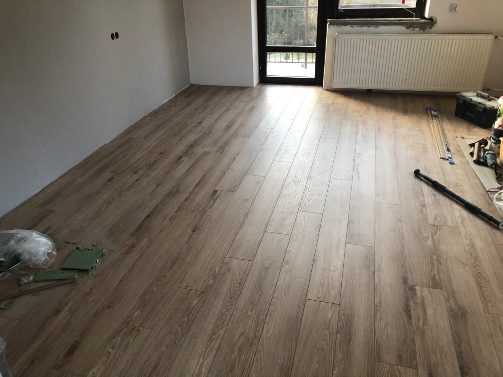 Nowo ułożone panele podłogowe w jasnym odcieniu drewna, widoczne narzędzia i akcesoria montażowe, ściany w szarym odcieniu, okno z widokiem na zieleń.