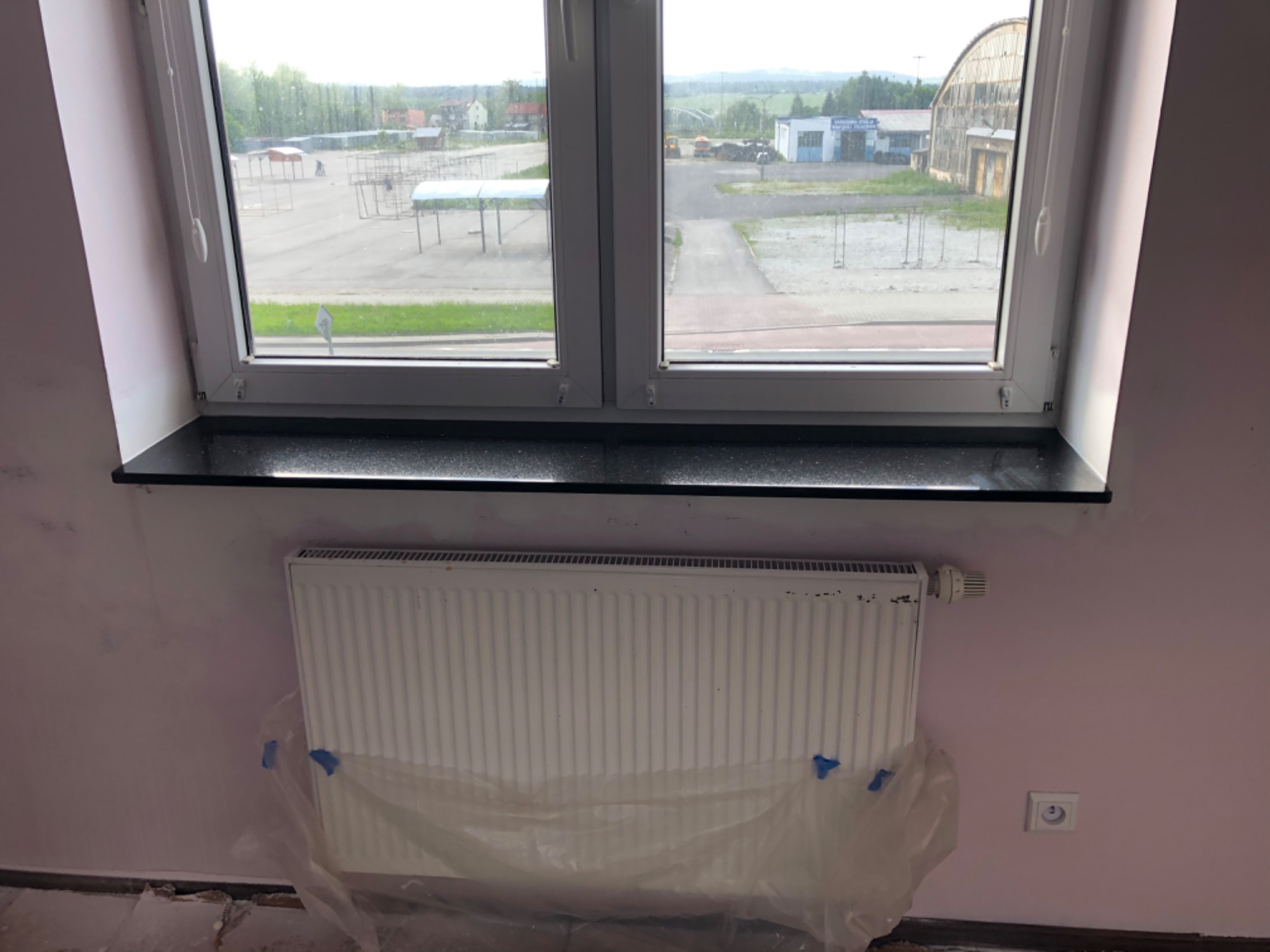 Nowy, czarny parapet z brokatem zamontowany pod białym oknem PCV; grzejnik zabezpieczony folią malarską, ściana pomalowana na jasny różowy kolor, widoczne ślady po remoncie na podłodze.
