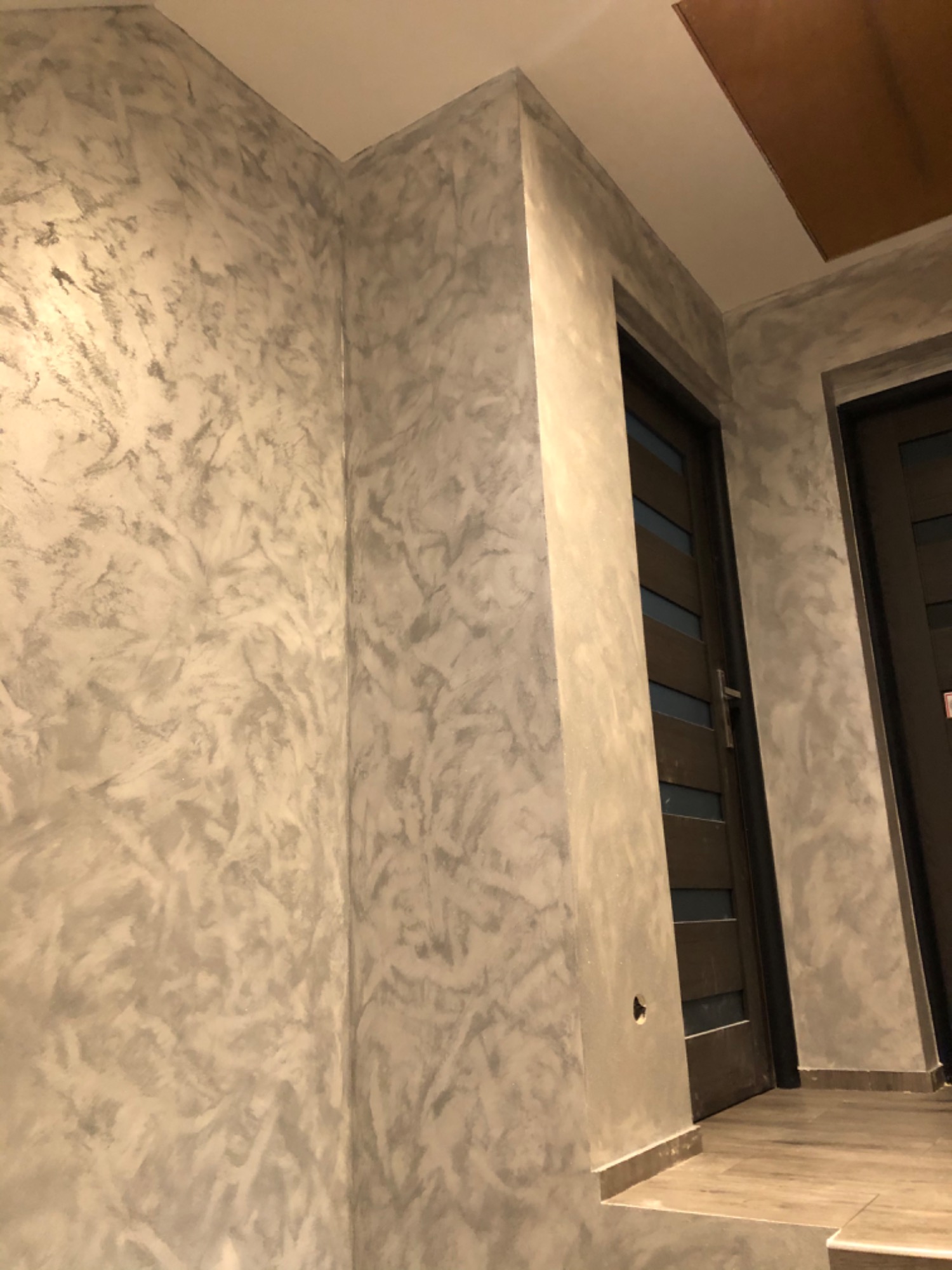 Ściana w wykończeniu dekoracyjnym imitującym beton, z widocznymi drzwiami z ciemnego drewna i szklanymi panelami, oświetlona ciepłym światłem.