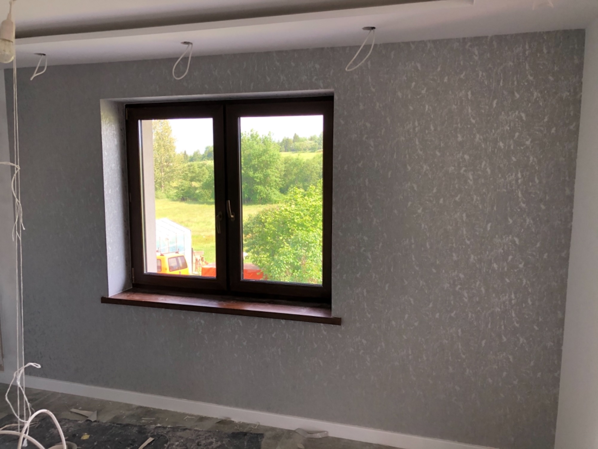 Ściana pokryta szarą tapetą ze wzorem, widok na okno z brązową ramą i parapetem, przez które widać zieleń drzew i żółty pojazd, na suficie widoczne wystające kable po lampie.