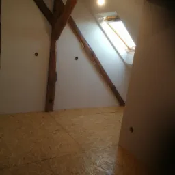 Poddasze w trakcie adaptacji: widoczne drewniane belki konstrukcyjne, płyty OSB na podłodze, okno dachowe i surowa ściana z przygotowanymi punktami elektrycznymi.