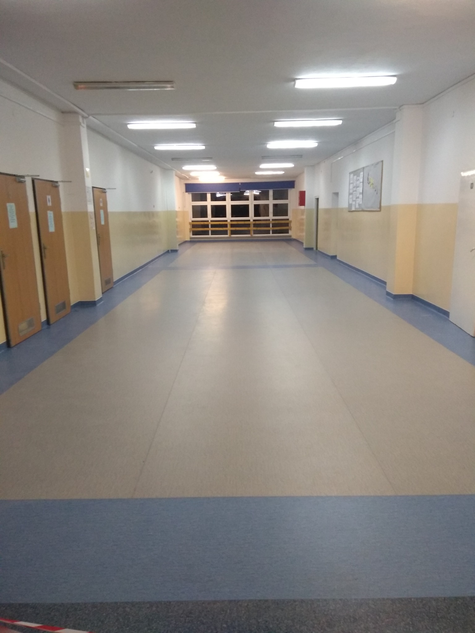 Długi korytarz w budynku użyteczności publicznej z jasną wykładziną podłogową z niebieskim pasem obwodowym, oświetlony szeregiem lamp sufitowych.