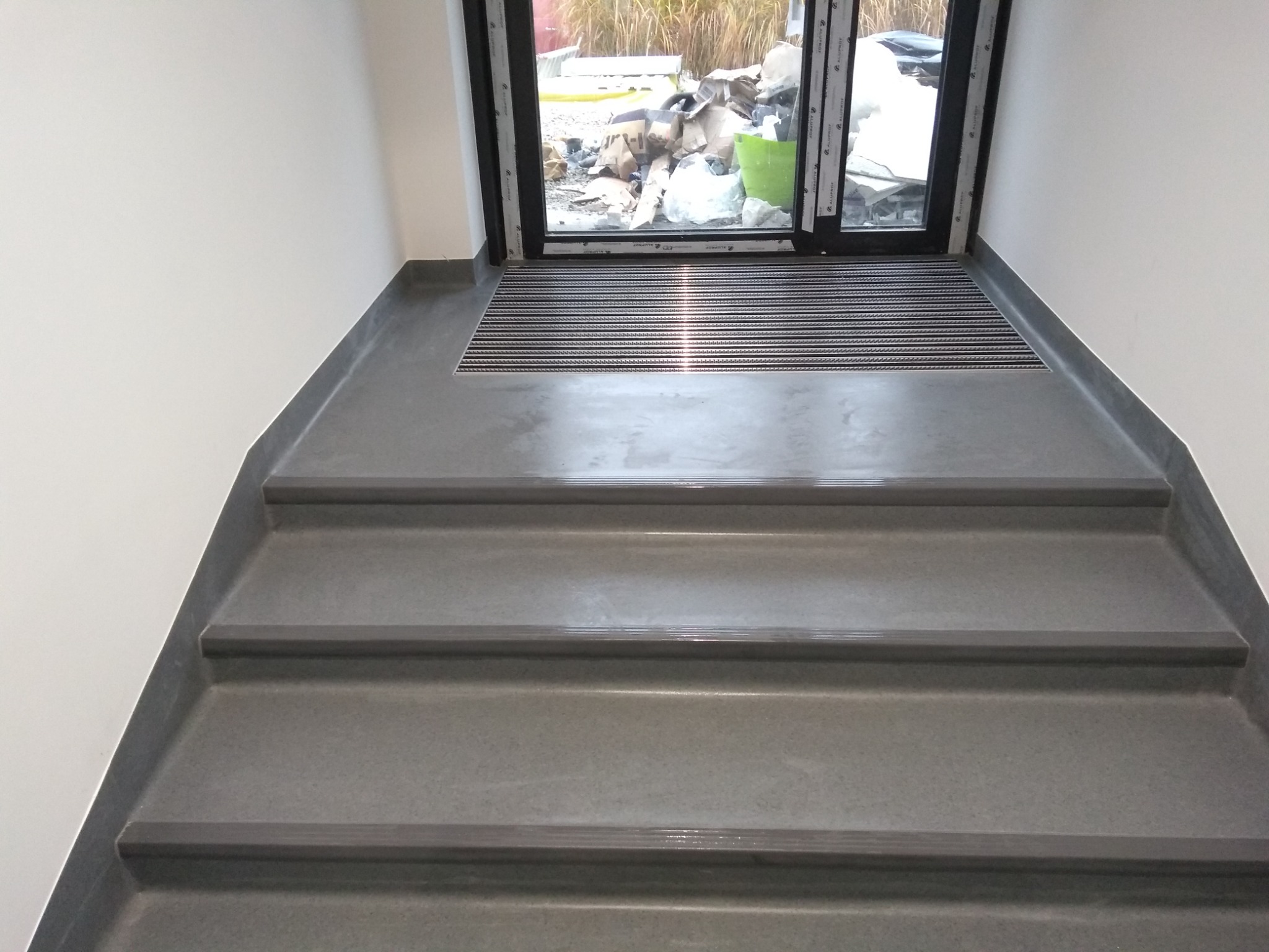 Szare schody pokryte wykładziną prowadzące do wejścia z wycieraczką; za oknem widoczne materiały budowlane.