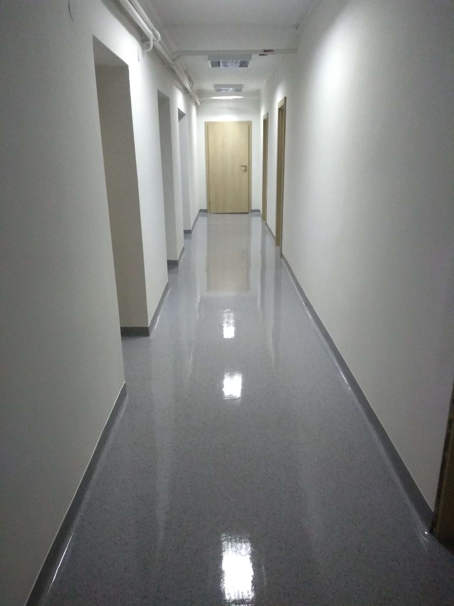 Długi korytarz z szarą, błyszczącą posadzką i białymi ścianami, widoczne drzwi z jasnego drewna na końcu korytarza.