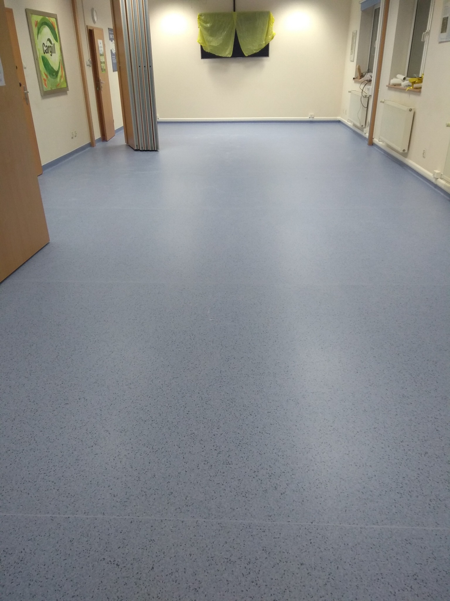 Jasnoszara wykładzina PCV z drobnym wzorem w dużej sali, widoczne drzwi i grzejniki.