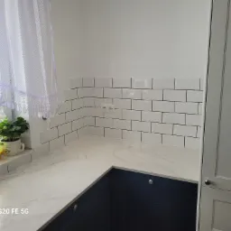 Kącik kuchenny z białymi, prostokątnymi płytkami ułożonymi w cegiełkę, blatem z białego marmuru, ciemnoniebieskimi szafkami i białymi szafkami po prawej stronie, przy oknie firanka z delikatną...