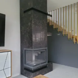 Obudowa kominka z ciemnego marmuru z metalowym wkładem, widok na schody z drewnianymi stopniami i złotymi barierkami, biała podłoga z delikatnym wzorem.