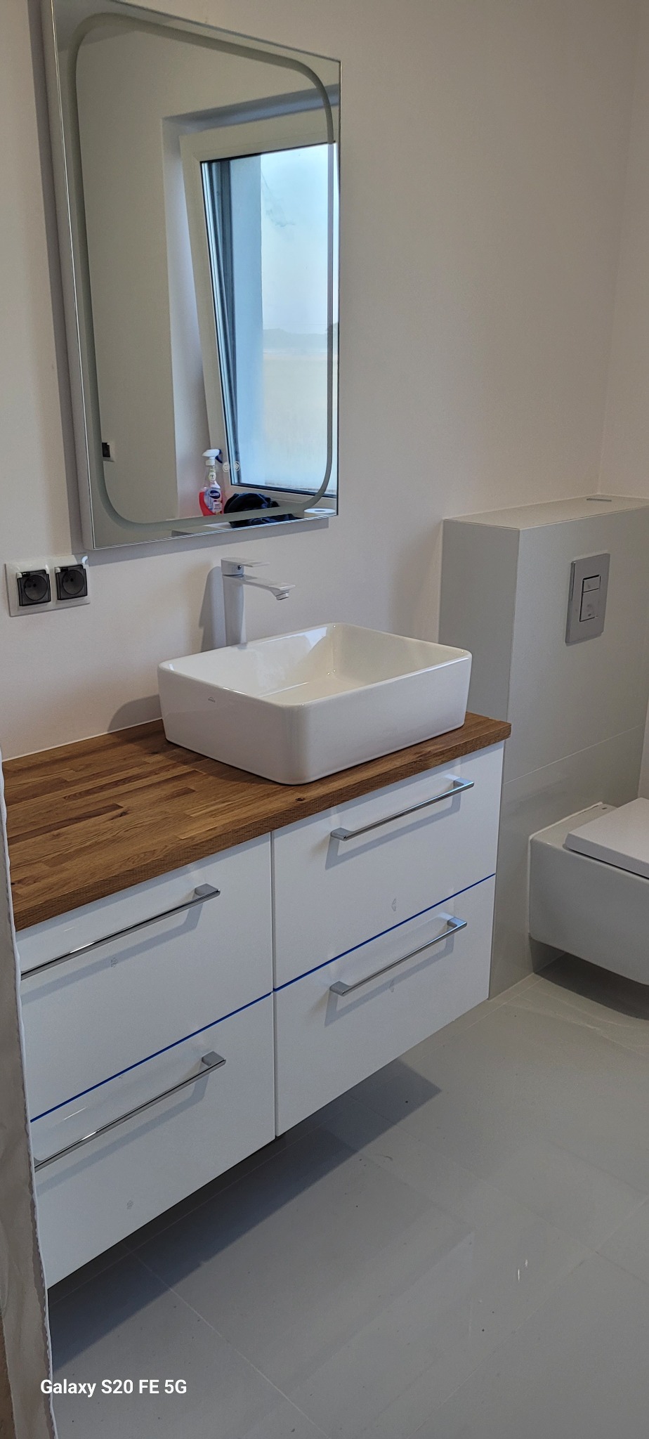 Nowoczesna łazienka z białą, prostokątną umywalką nablatową, drewnianym blatem i szafkami z podłużnymi uchwytami. Nad umywalką wisi podświetlane lustro z zaokrąglonymi rogami, w którym odbija się...