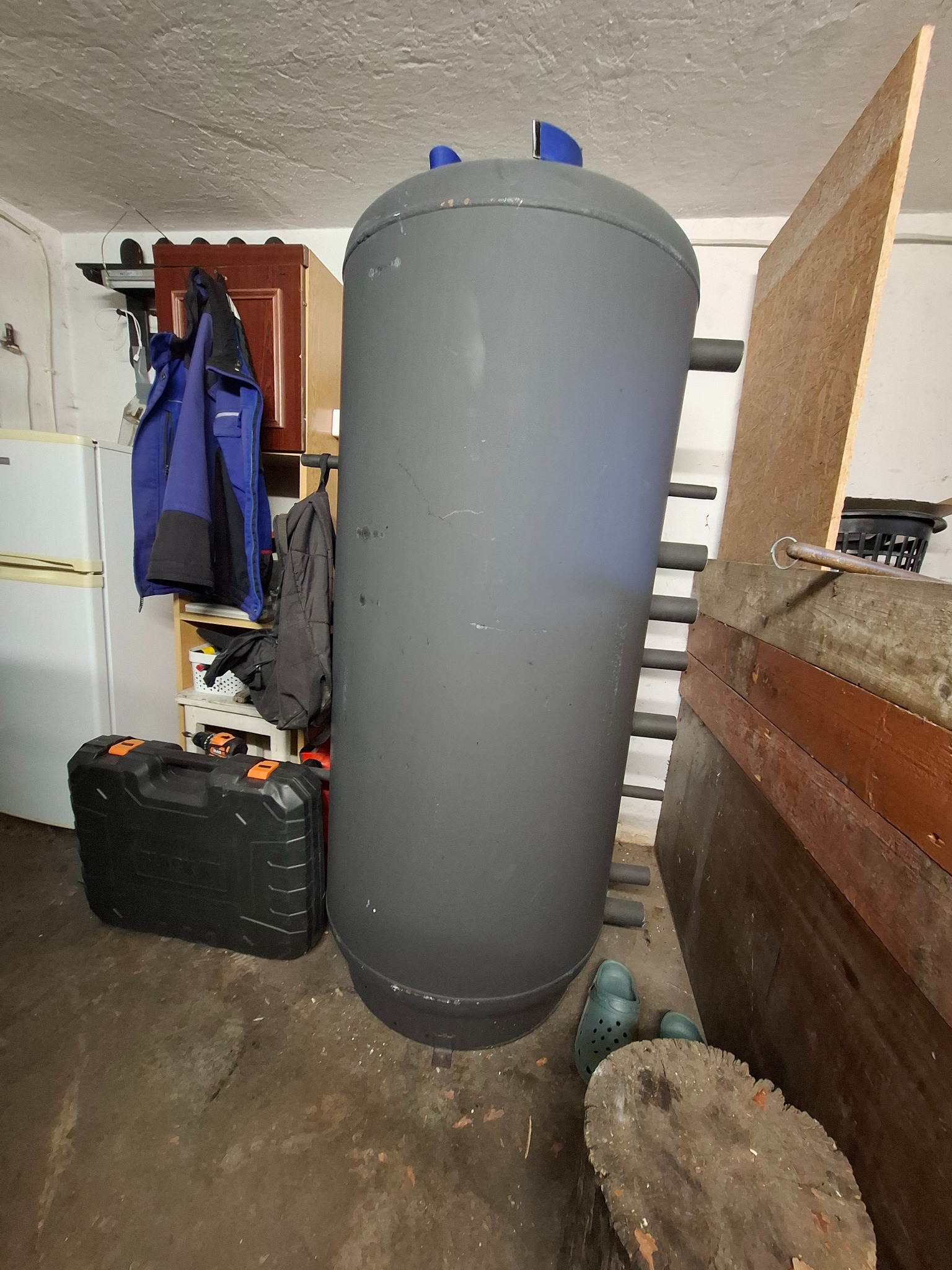 Szary, pionowy zbiornik z niebieskimi elementami u góry, obok skrzynka narzędziowa, drewniane deski i zielone chodaki w pomieszczeniu gospodarczym.