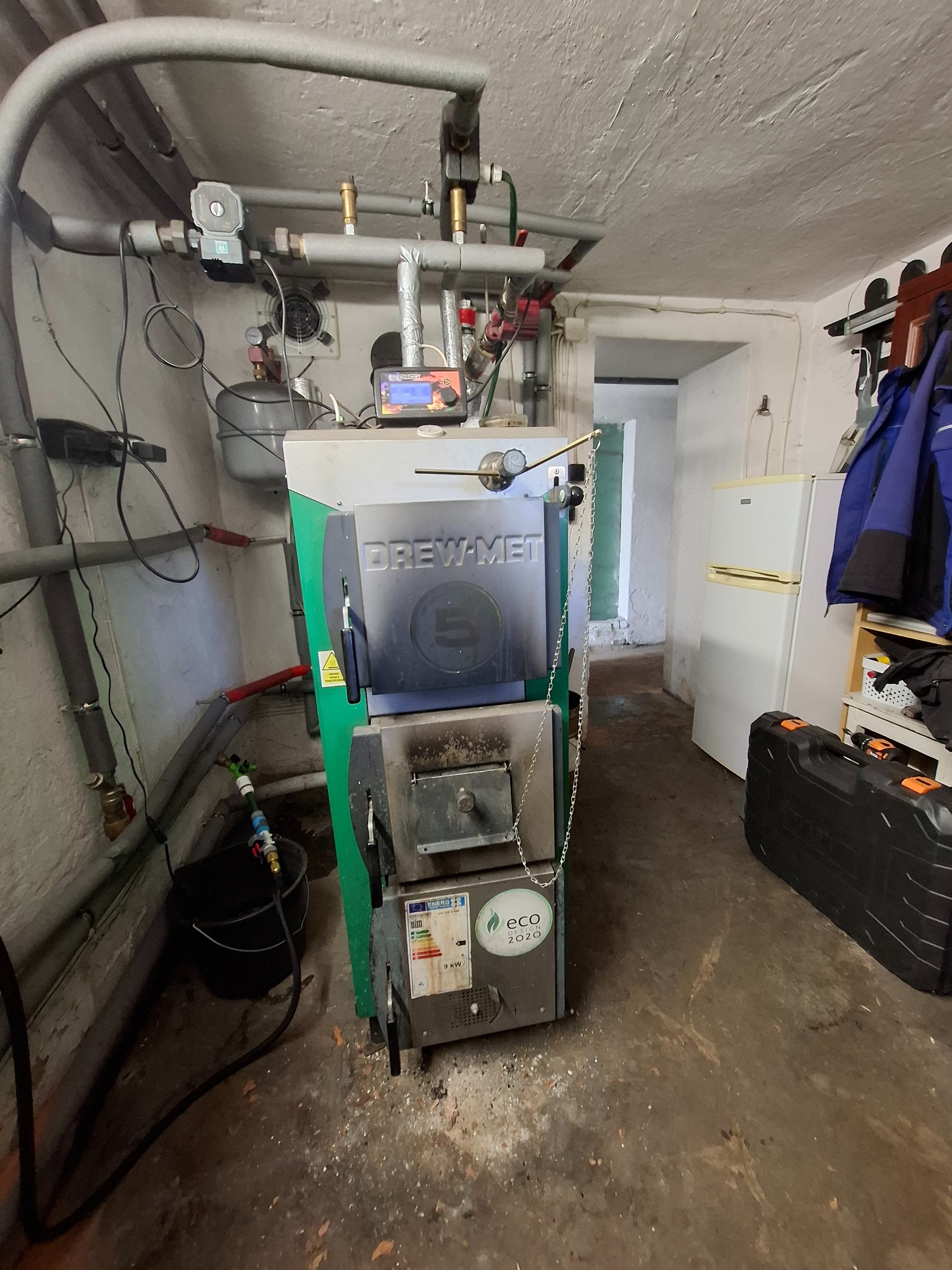 Kocioł grzewczy Drew-Met w piwnicy, widoczne rury, instalacja, regulator temperatury i czarny kufer. Biała ściana i lodówka w tle.