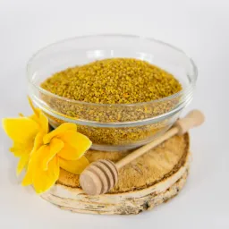 Szklana miseczka wypełniona pyłkiem kwiatowym na drewnianym plastrze z korą brzozy, obok żółty kwiat i drewniany nabierak do miodu.