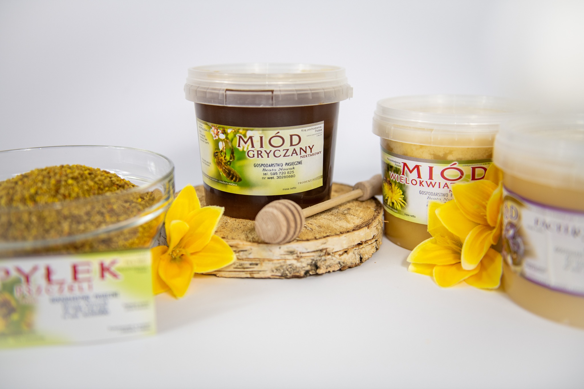 Słoiki z miodem gryczanym i wielokwiatowym z Wągrowca, pyłek pszczeli w szklanej miseczce, drewniany nabierak do miodu i żółte kwiaty na białym tle.
