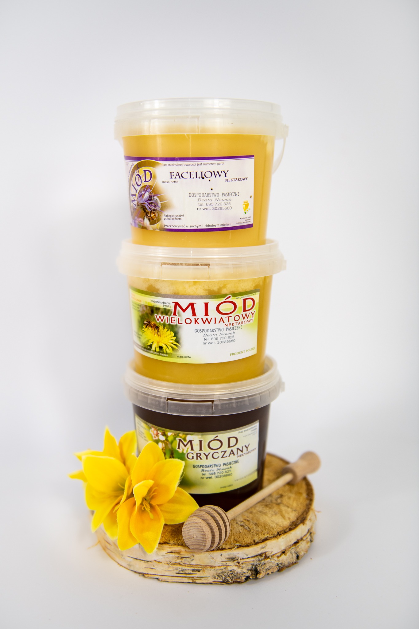 Wieża z trzech plastikowych pojemników z różnymi rodzajami miodu (faceliowy, wielokwiatowy, gryczany), ułożonych na kawałku drewna z korą, obok leży drewniany nabierak do miodu i żółty kwiat.