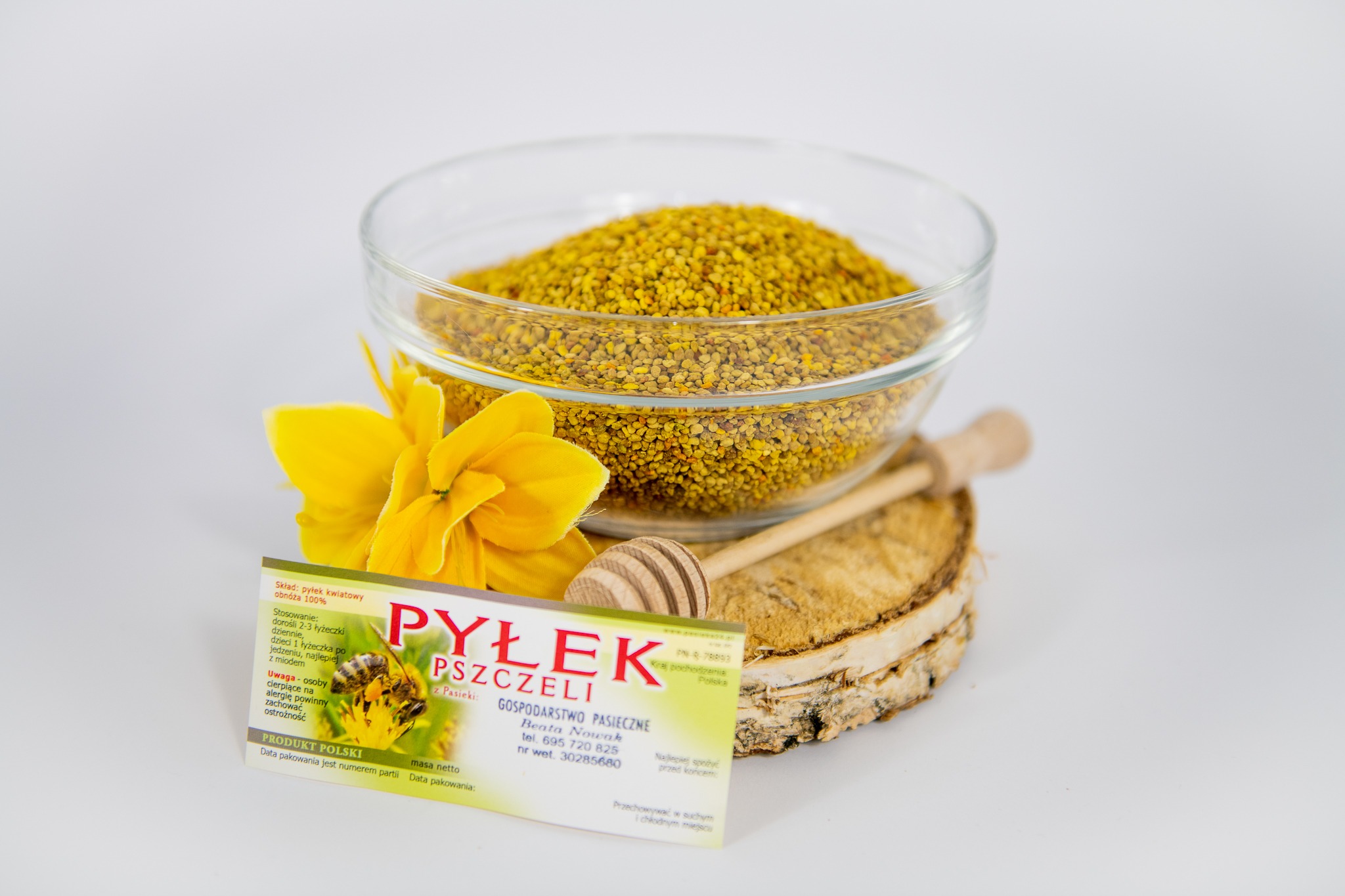 Pyłek pszczeli w szklanej miseczce, drewniana łyżeczka, plaster drewna i ulotka informacyjna z wizerunkiem pszczoły na kwiatku, całość na białym tle.