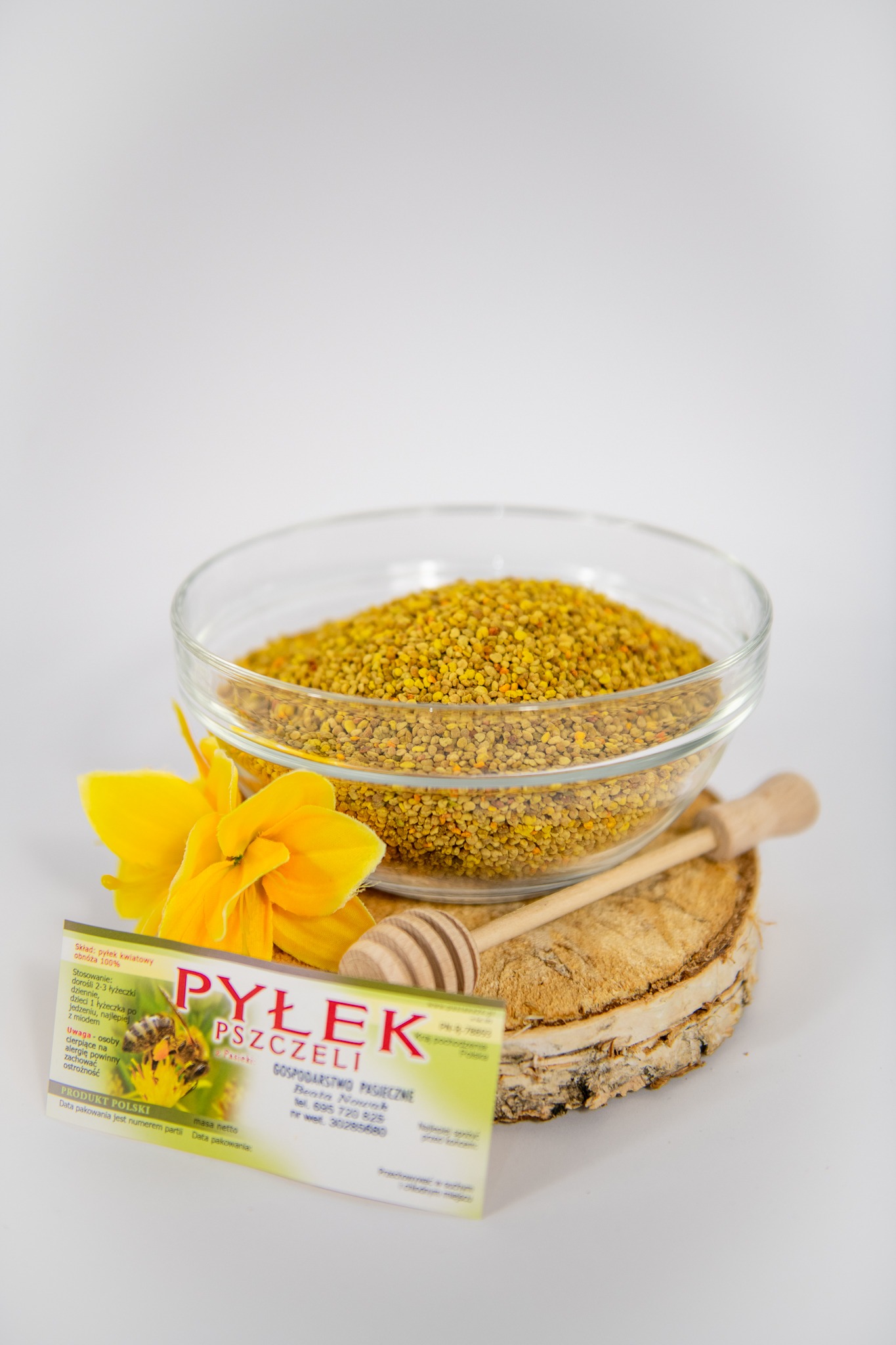 Szklana miseczka wypełniona pyłkiem pszczelim, ułożona na drewnianym plastrze obok żółtego kwiatu i drewnianej łyżeczki do nabierania, obok ulotka informacyjna produktu
