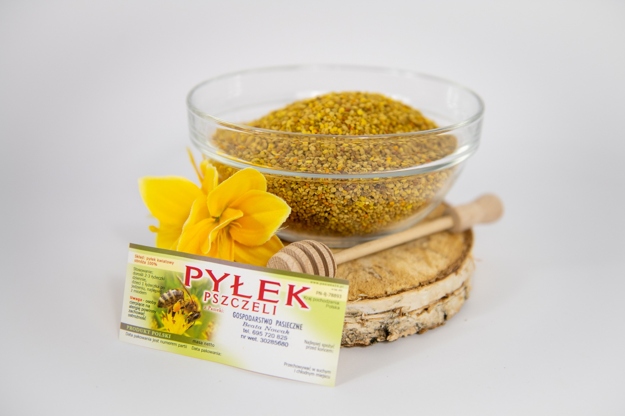 Pyłek pszczeli w szklanej miseczce, obok żółty kwiat i wizytówka pasieki na drewnianym plastrze, z łyżeczką do miodu, na białym tle.
