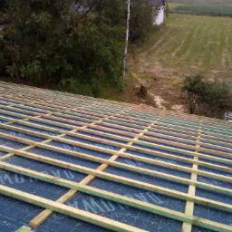 W trakcie budowy dachu: widok na konstrukcję drewnianą z łat i kontrłat ułożonych na niebieskiej membranie dachowej, w tle zieleń drzew i pole.