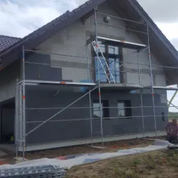 Dom jednorodzinny w trakcie ocieplania styropianem, widoczne rusztowanie i częściowo ocieplona ściana, na ziemi leżą elementy rusztowania i folia ochronna.