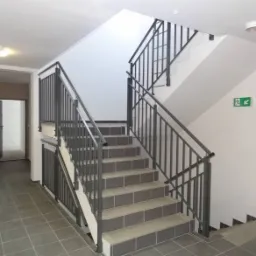 Klatka schodowa w budynku mieszkalnym z szarymi płytkami na podłodze i schodach, metalowe poręcze, białe ściany z zielonym znakiem ewakuacyjnym, widoczne drzwi w tle.