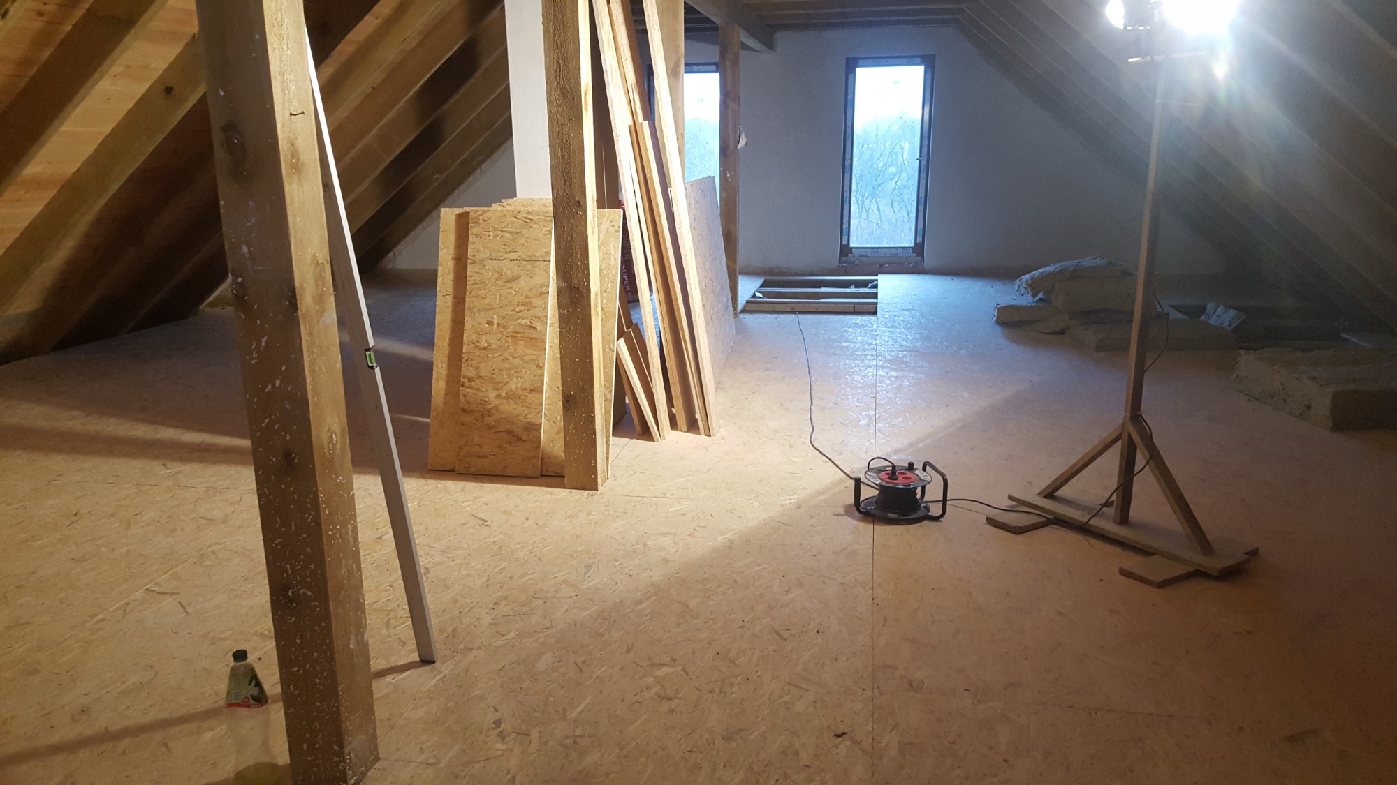 Poddasze w trakcie ocieplania: widoczne płyty OSB na podłodze, wełna mineralna, drewniane belki, poziomica oparta o filar oraz lampa budowlana na trójnogu oświetlająca przestrzeń.