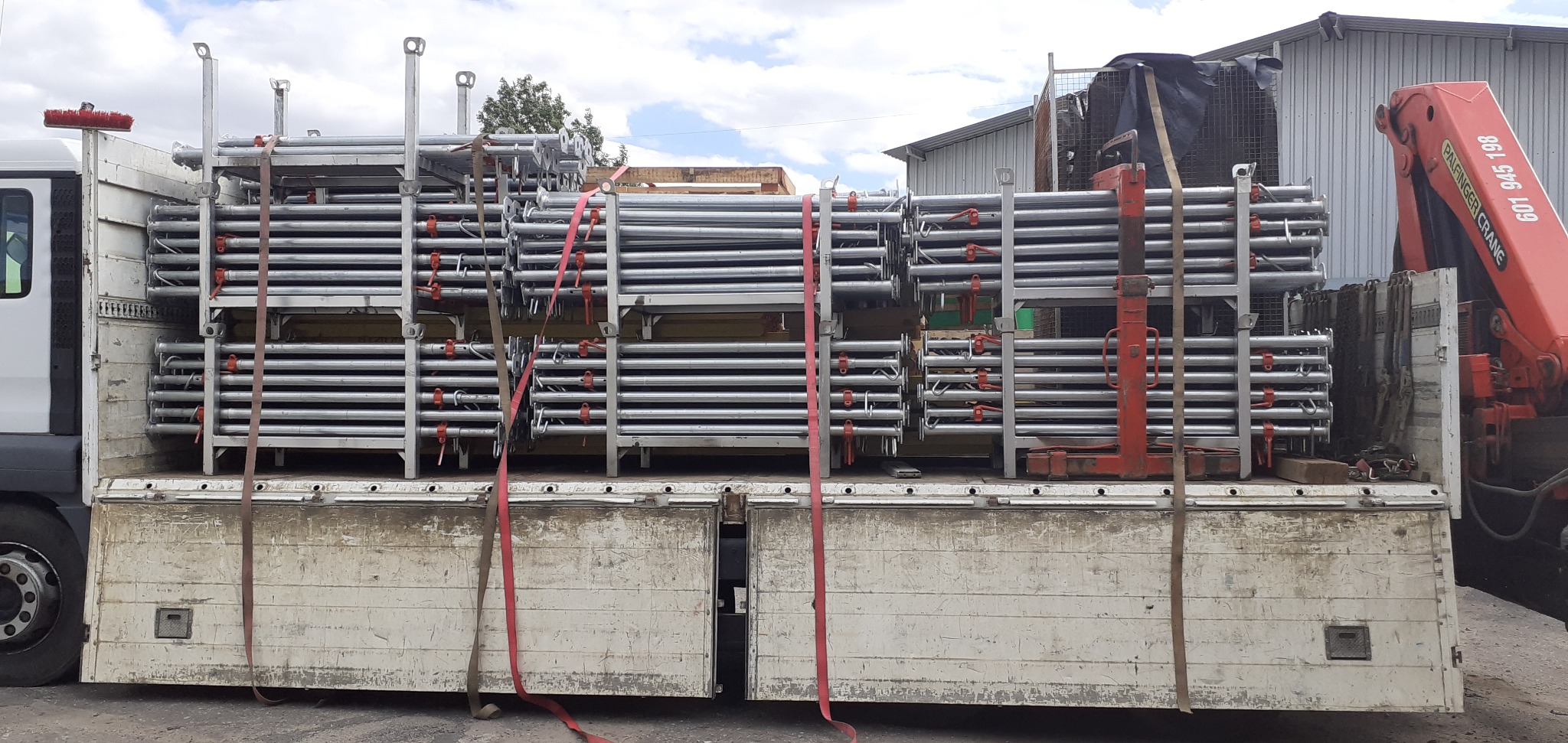 Załadunek metalowych rusztowań budowlanych na skrzynię samochodu dostawczego z dźwigiem hydraulicznym w tle.