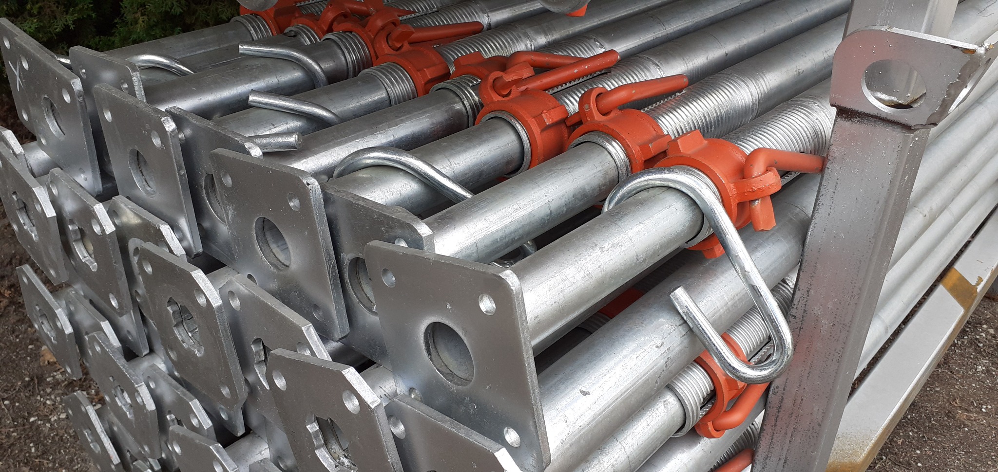 Stos ocynkowanych podpór budowlanych z pomarańczowymi zaciskami, ułożonych równo, widoczne kwadratowe podstawy z otworami i gwintowane sekcje.