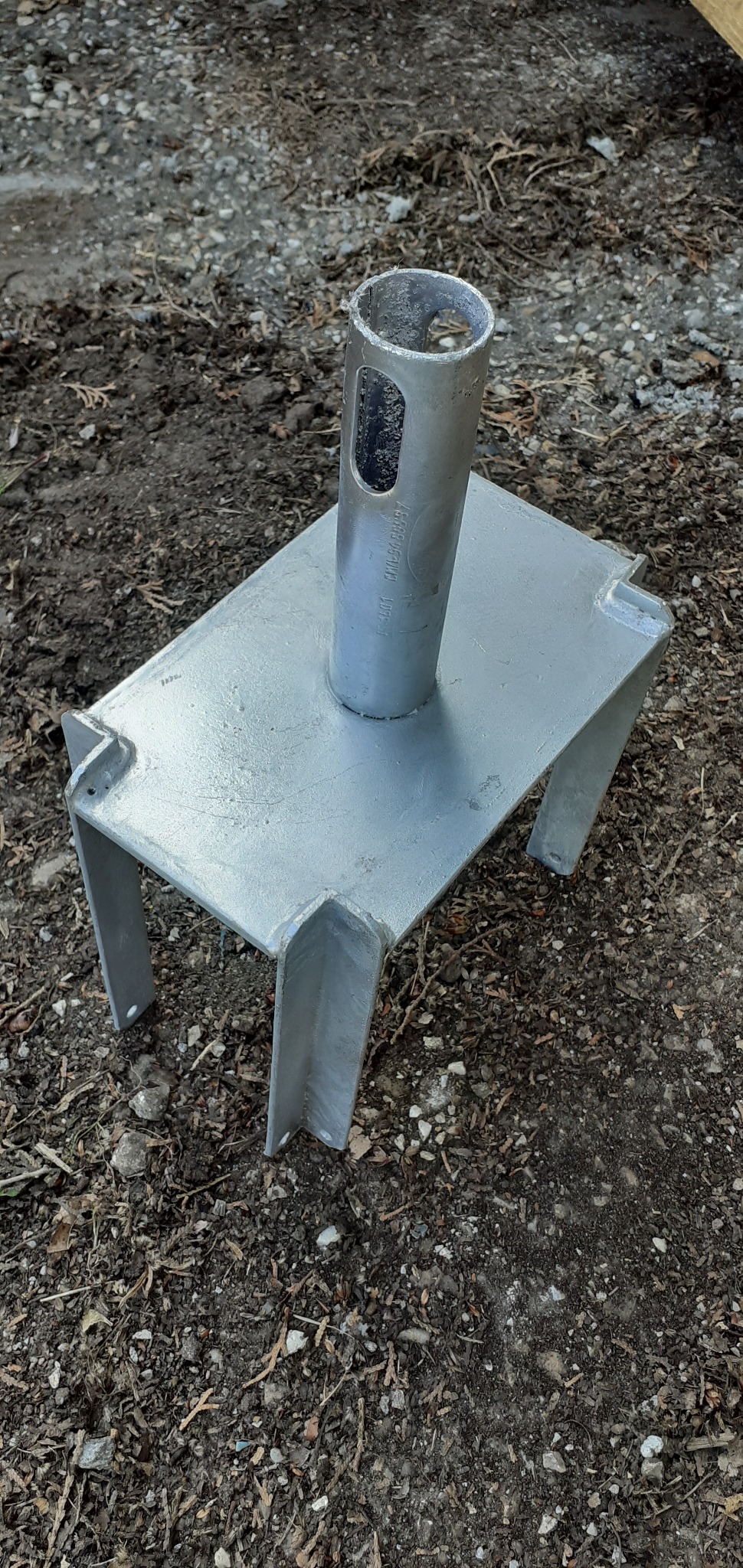 Metalowa podstawa słupa z czterema nogami, ustawiona na ziemi z drobnymi kamieniami i resztkami roślin.