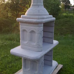 Niedokończona, dwupoziomowa konstrukcja z bloczków betonowych imitująca grill ogrodowy z dekoracyjnymi elementami, ustawiona na palecie na trawniku w ogrodzie, w tle drzewa w popołudniowym słońcu.