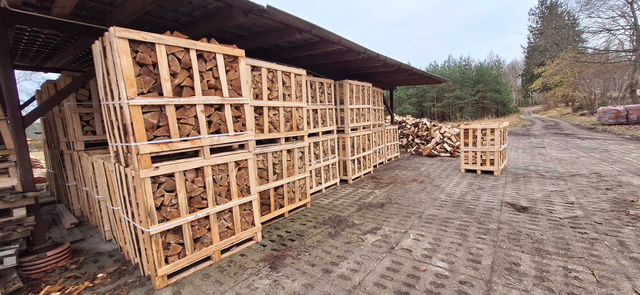 Ułożone w paletach pod zadaszeniem, gotowe do transportu pocięte kawałki drewna opałowego, na zewnątrz sterta drewna i leśna droga.