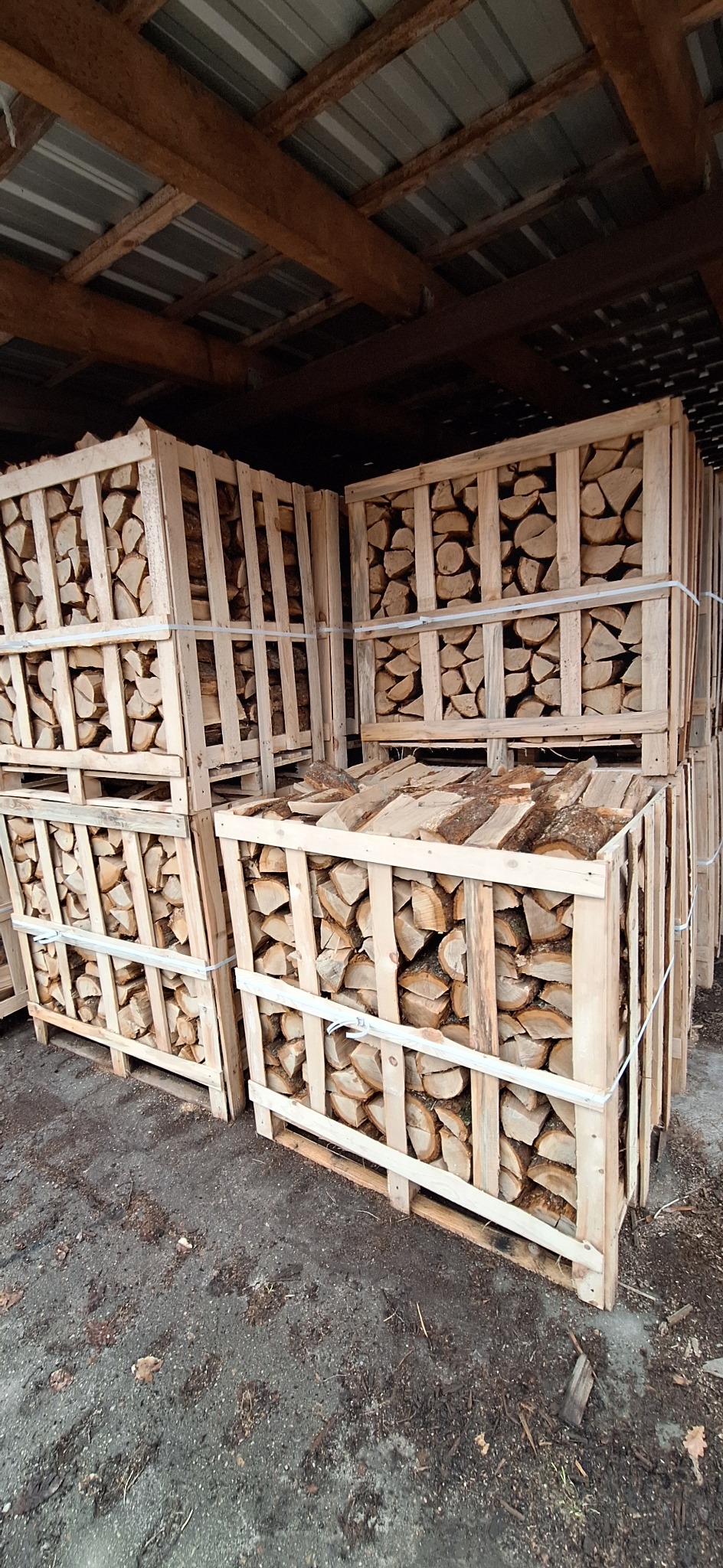 Stos palet z drewnem kominkowym pod zadaszeniem, widoczne nierówne końcówki polan i szare podłoże.