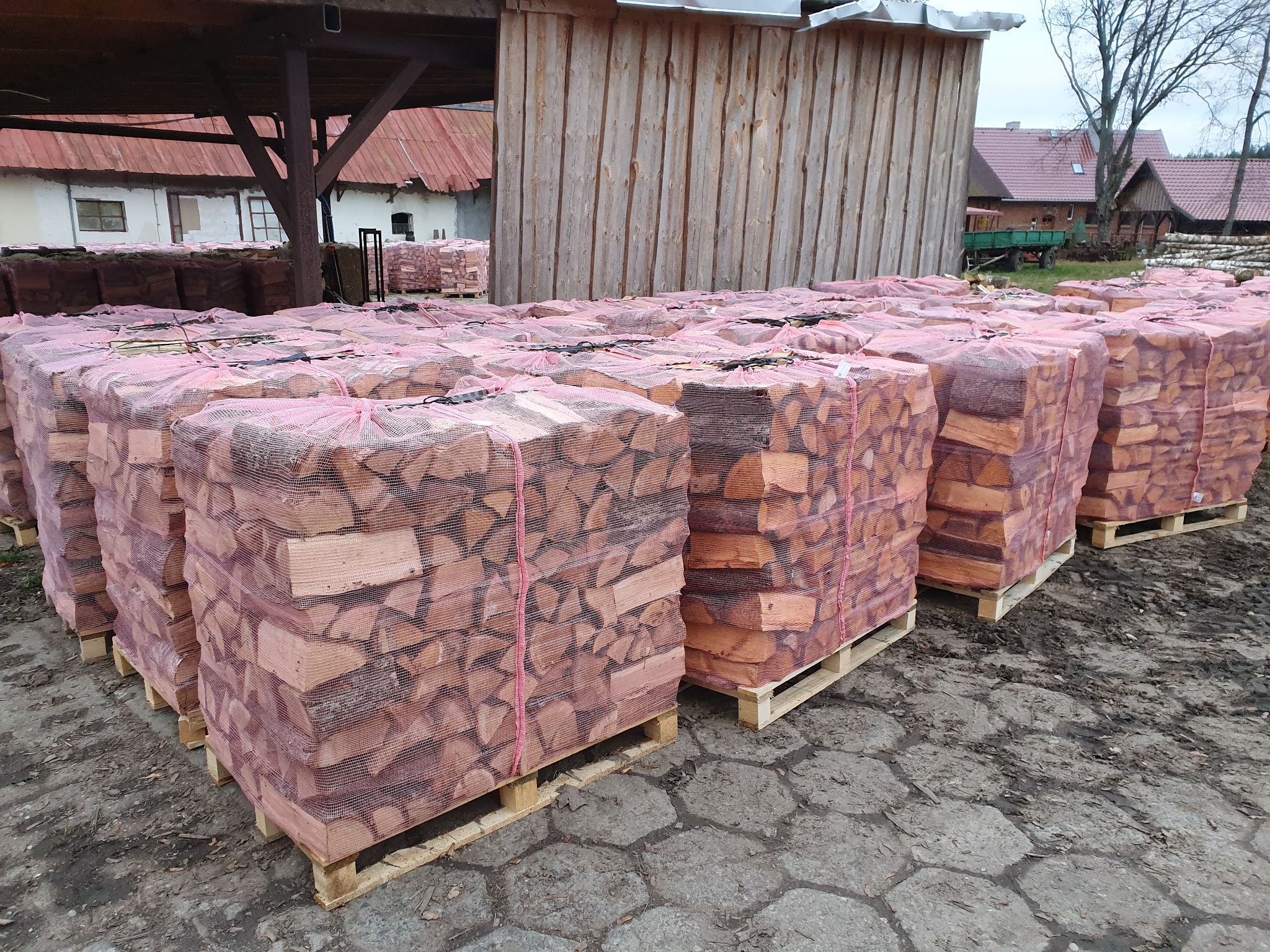 Ułożone w rzędach, pocięte i spakowane drewno kominkowe na paletach, zabezpieczone siatką w kolorze różowym, na betonowym placu w tle zabudowania gospodarcze.