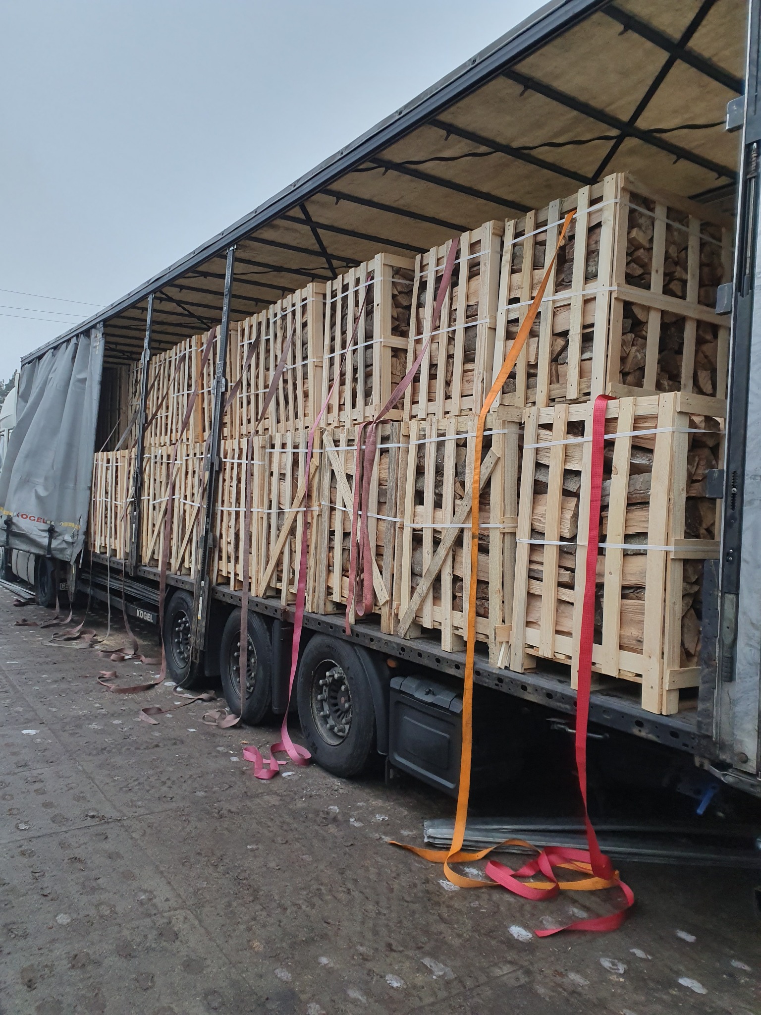 Ciężarówka z otwartą naczepą załadowana paletami z pociętym drewnem opałowym, zabezpieczona pasami transportowymi.