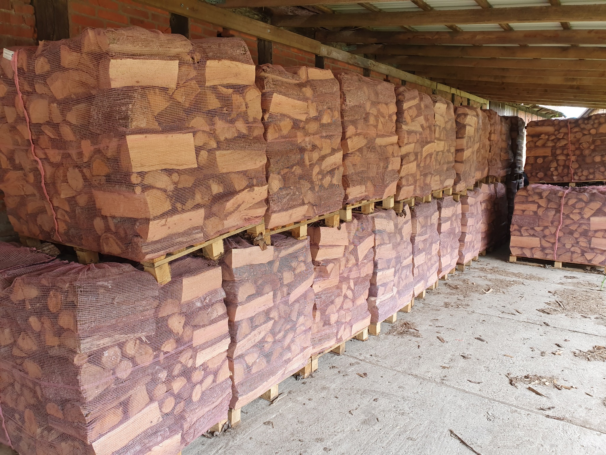 Rzędami ułożone worki z pociętym drewnem na paletach, składowane pod zadaszeniem z ceglaną ścianą w tle.