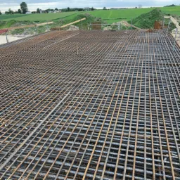 Siatka zbrojeniowa dwuwarstwowa, przygotowana pod wylewkę fundamentów, widok z góry na tle zielonych pól i pochmurnego nieba.