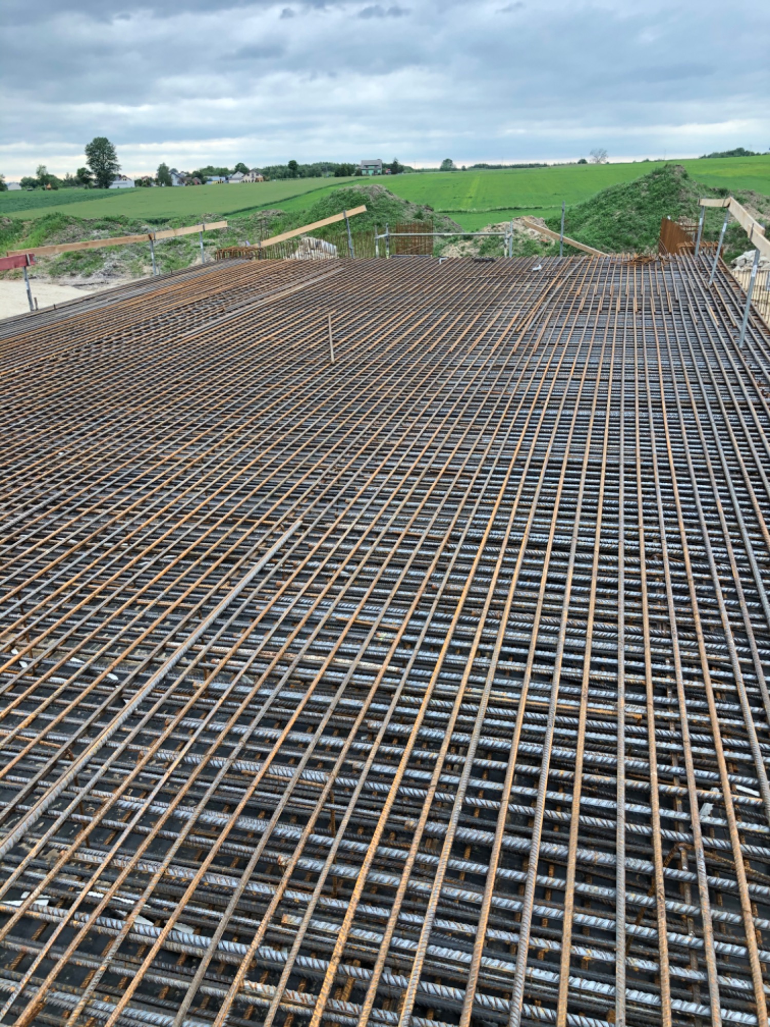 Siatka zbrojeniowa dwuwarstwowa, przygotowana pod wylewkę fundamentów, widok z góry na tle zielonych pól i pochmurnego nieba.