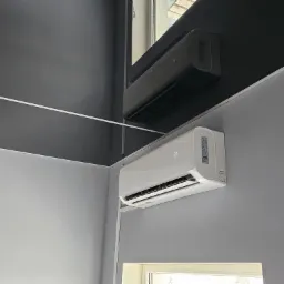 Dwa klimatyzatory ścienne: biały model AUX z wyświetlaczem temperatury i czarny, zamontowane w rogu pomieszczenia o szarych ścianach i suficie.