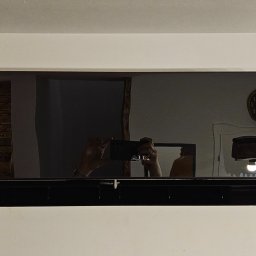 Carmet - Czarny klimatyzator ścienny z wyświetlaczem temperatury, widoczne odbicie wnętrza z zegarem i lampą, osoba robiąca zdjęcie w odbiciu.