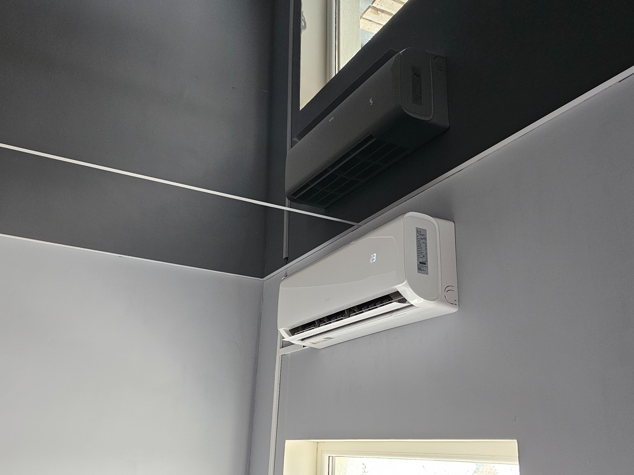 Dwa klimatyzatory ścienne: biały model AUX z wyświetlaczem temperatury i czarny, zamontowane w rogu pomieszczenia o szarych ścianach i suficie.