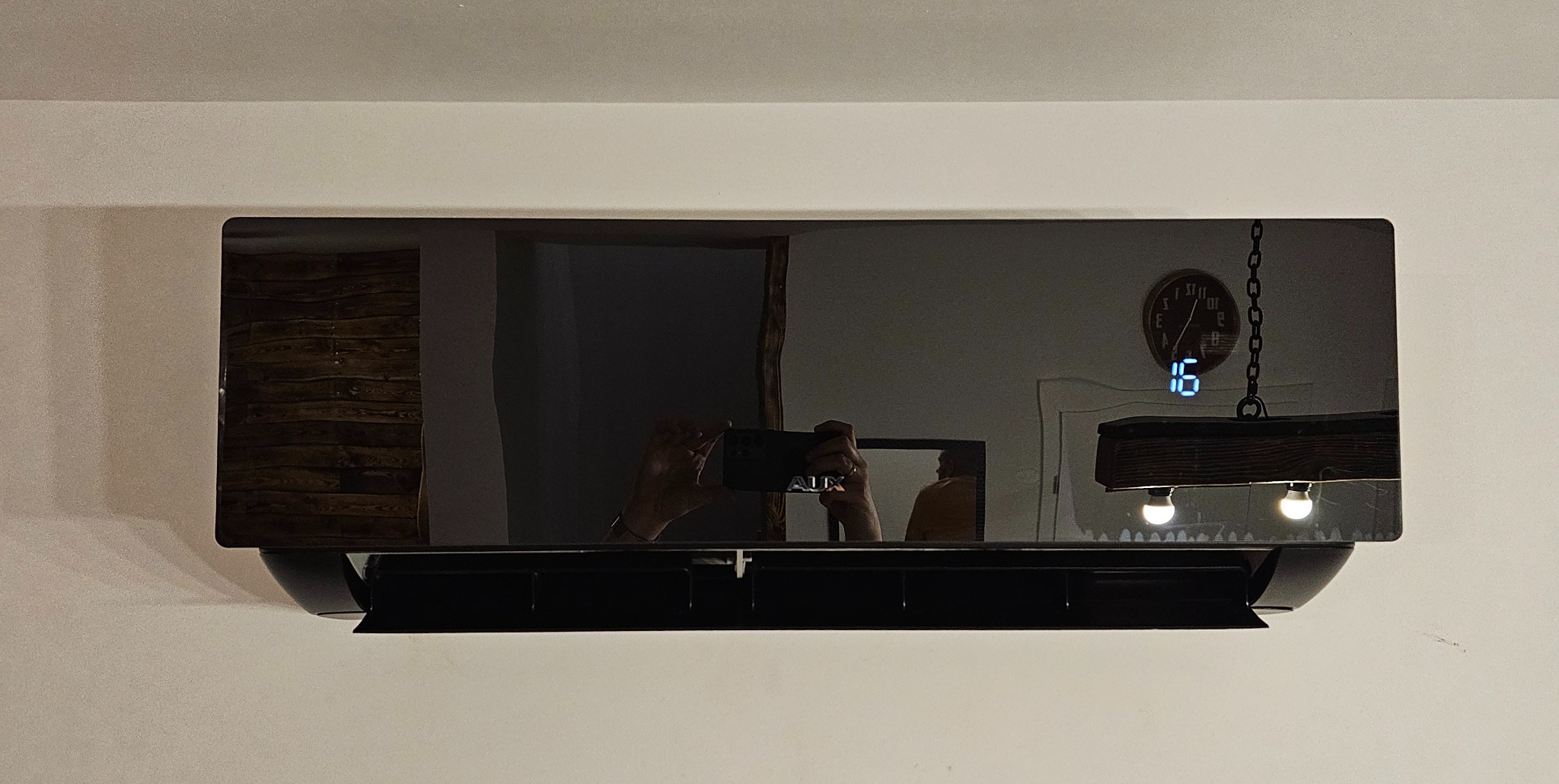 Czarny klimatyzator ścienny z wyświetlaczem temperatury, widoczne odbicie wnętrza z zegarem i lampą, osoba robiąca zdjęcie w odbiciu.