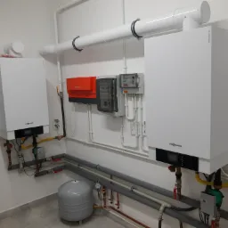 Dwa białe piece grzewcze Viessmann z podłączonymi rurami, zaworami i osprzętem, zamontowane w kotłowni z szarym zbiornikiem wyrównawczym i białą wentylacją.