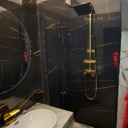 Elegancka łazienka z czarnymi płytkami imitującymi marmur z złotymi żyłami, kabina prysznicowa typu walk-in ze złotą armaturą, umywalka nablatowa na szafce z marmurowym blatem i złotymi akcentami...