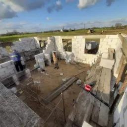 Wznoszenie ścian parterowego domu z bloczków betonu komórkowego. Widoczne rusztowania, pracownicy w trakcie murowania, niezadaszona konstrukcja oraz krajobraz wiejski w tle pod błękitnym niebem...