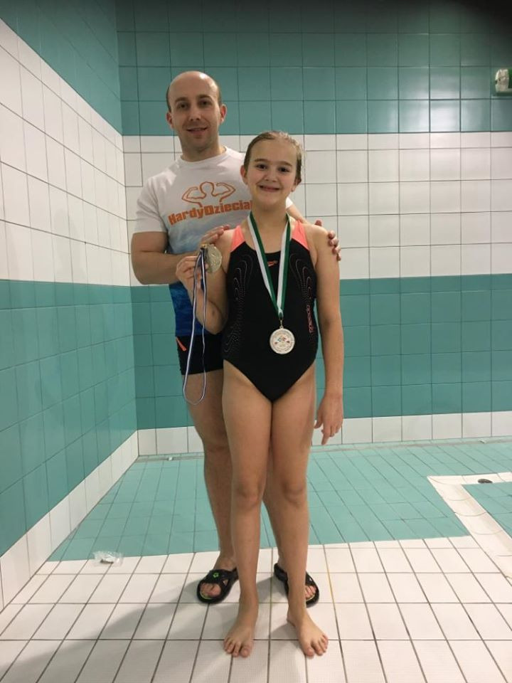 Dziewczynka w stroju kąpielowym z medalami, pozująca z trenerem na tle basenu z biało-turkusowymi kafelkami.