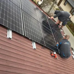 Montaż paneli słonecznych na dachu z blachy trapezowej przez dwóch instalatorów, widoczny wkrętak akumulatorowy i elementy montażowe.